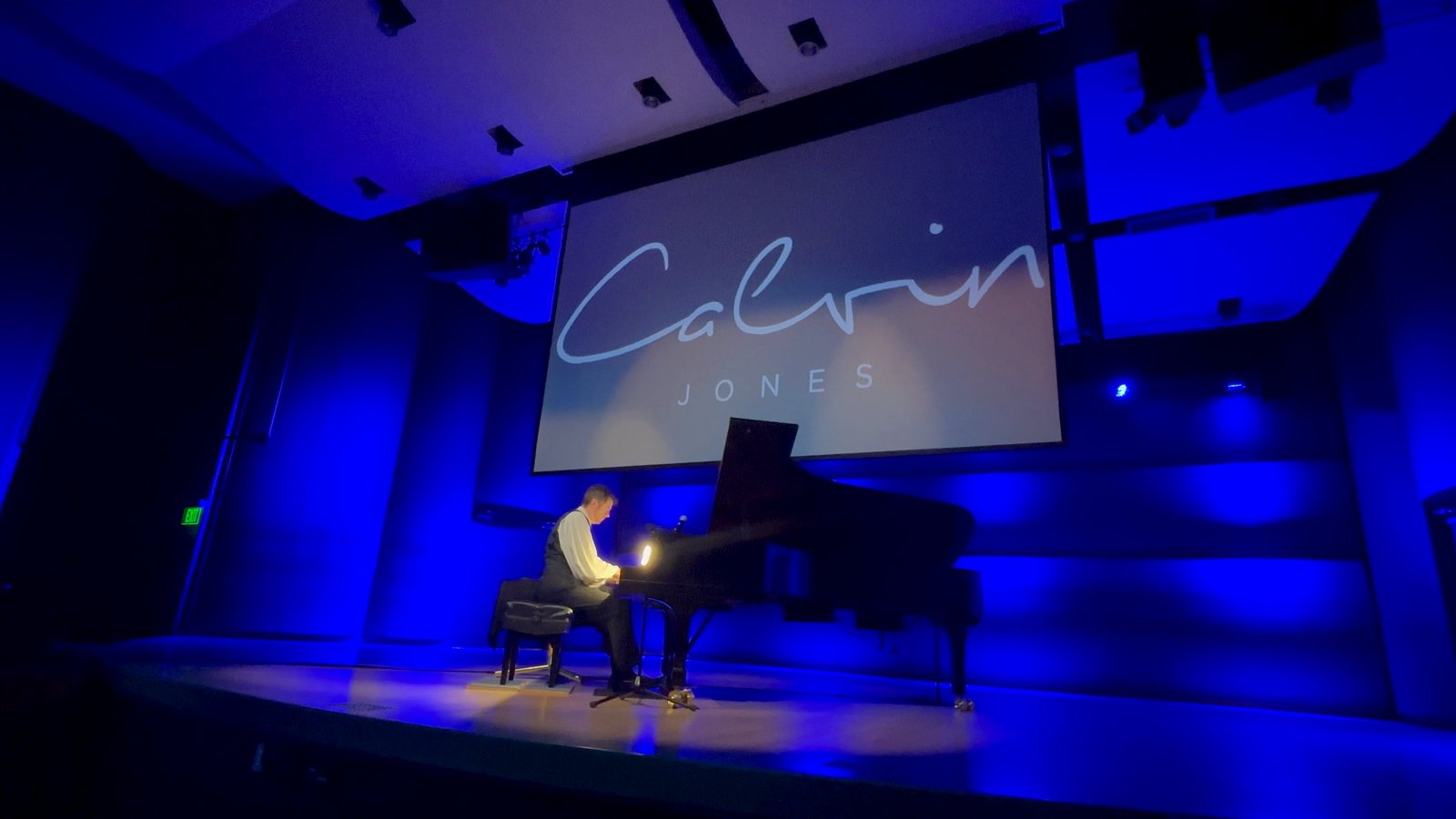 Calvin Jones at the grand piano beneath a Calvin Jones title screen, bathed in blue stage light
